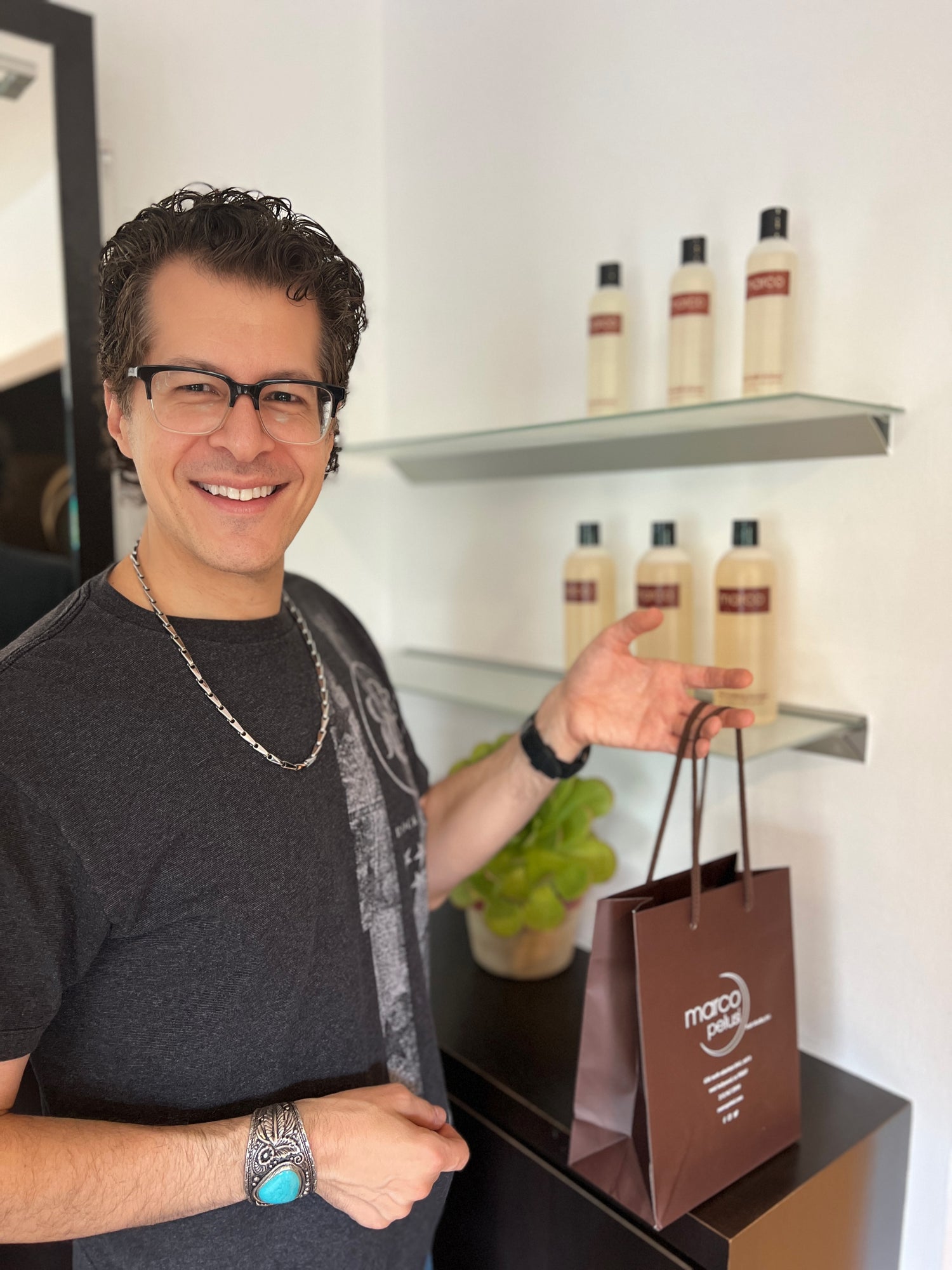Marco Pelusi holding a brown "Marco Pelusi Hair Studio" shopping bag with a shelf of marco product bottles in the background